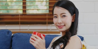 近距离的年轻亚洲女人拿着红色的咖啡杯，吸入和微笑时看着镜头，而坐在家里客厅的沙发背景，人们的饮食生活方式