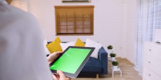 女人用绿屏平板电脑应用来控制家里客厅的空调。智能家居自动化控制概念