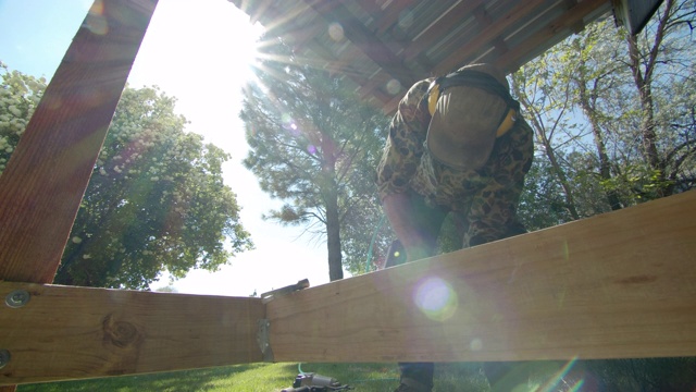 一个40多岁的白人杂工戴着帽子、太阳镜、听力保护和护腿，在一个阳光明媚的日子里，在一个居民区的户外搭建甲板时，用气动钉枪把金属角支架固定在木板上