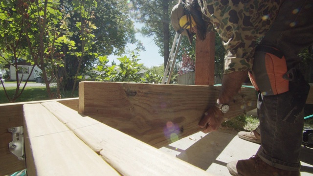 一个40多岁的白种杂工，戴着帽子、太阳镜、听力保护和护腿，在一个居民区的户外阳光下建造一个甲板时，把一块木板放在一起，然后用气动钉枪把木板固定在一起