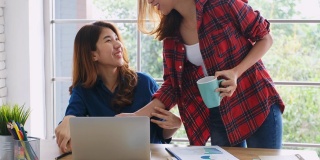两个年轻的亚洲女性工作与笔记本电脑和喝咖啡在家庭办公室与幸福，办公室休闲的生活方式