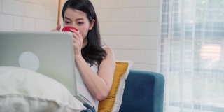 年轻的亚洲自由职业女性在家里的客厅休息时，用笔记本电脑查看社交媒体和喝咖啡。居家女性的生活方式概念。