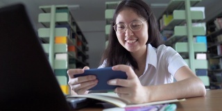 亚洲大学生女孩读书，工作和网上购物与笔记本电脑和智能手机在公共图书馆做她的家庭作业