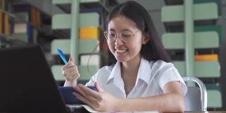 亚洲大学生女孩读书和工作用笔记本电脑和智能手机做她的家庭作业在公共图书馆
