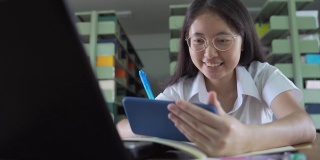 亚洲大学生女孩读书，工作和网上购物与笔记本电脑和智能手机在公共图书馆做她的家庭作业