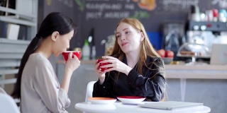 两名年轻女同事在咖啡馆边喝咖啡边聊天的中镜头。亚裔和白人女商人坐在桌边聊天