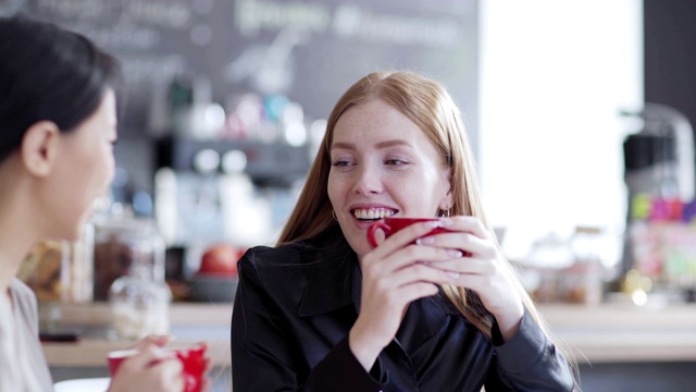 右图是两个年轻女人在咖啡馆喝茶或喝咖啡，愉快地聊天。红发白人女性聊天与分散的亚洲朋友