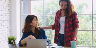 两个年轻的亚洲女性工作与笔记本电脑和喝咖啡在家庭办公室与幸福，办公室休闲的生活方式