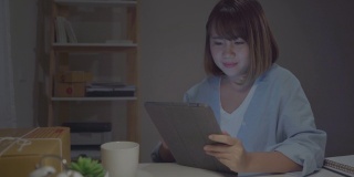 亚洲年轻的女企业家企业主中小企业在线检查产品库存，保存到平板电脑和喝咖啡在家里工作到深夜。小企业主在家办公的概念。