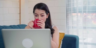 年轻的亚洲自由职业女性在家里的客厅休息时，用笔记本电脑查看社交媒体和喝咖啡。居家女性的生活方式概念。