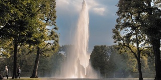 德国汉诺威。喷水池从放在地上的碗里喷出的巨大的高喷水池以公园里的绿树为背景。泌尿系统疾病和前列腺炎的治疗概念。慢动作