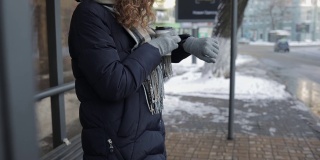 在冬天的一天，一个陌生的年轻女子穿着暖和的衣服在车站等车，喝着咖啡，看着手表。
