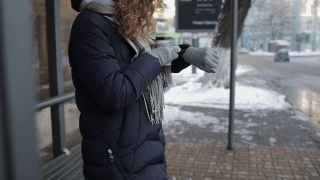 在冬天的一天，一个陌生的年轻女子穿着暖和的衣服在车站等车，喝着咖啡，看着手表。视频素材模板下载