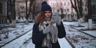 一名年轻女子用智能手机的相机当镜子涂口红，站在下雪的城市街道上。