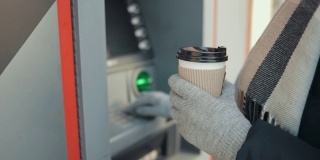 特写的年轻女子拿着咖啡和使用ATM在城市街道在冬天的一天。