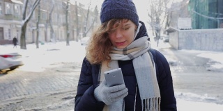 快乐的年轻女人戴着温暖的围巾和帽子在手机上打字和微笑在冬天在城市的道路附近的户外。