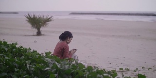 年轻女子用智能手机拍照，她正坐在沙滩上放松。