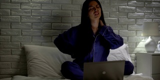 疲惫的女人按摩脖子，坐在床上工作时笔记本电脑不舒服