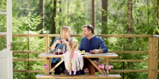 右图是三口之家围坐在乡间别墅门廊的木桌边喝茶，谈笑风生。父母高兴地听着他们可爱的小女儿的笑声