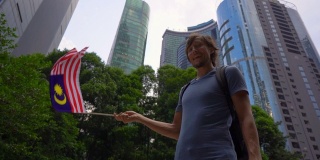 一名年轻男子手持智能手机挥舞着以摩天大楼为背景的马来西亚国旗的慢镜头。去马来西亚旅游的概念