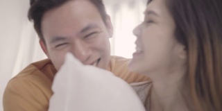 亚洲夫妇在卧室的床上玩枕头大战，妻子和丈夫在家里躺在床上享受有趣的时刻。夫妻在家里放松享受爱的时刻的概念。