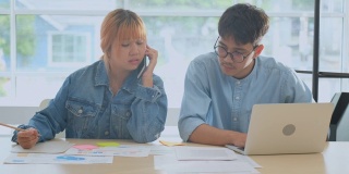 两个年轻的富有创造力的亚洲男人和女人谈论手机，同时工作在笔记本电脑和文书工作在放松，人们在办公室的生活方式