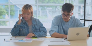两个年轻的富有创造力的亚洲男人和女人谈论手机，同时工作在笔记本电脑和文书工作在放松，人们在办公室的生活方式