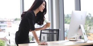 在办公室里，年轻的女商人用电脑站在办公桌前喝咖啡