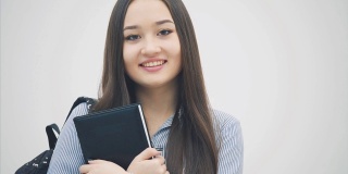 一名亚洲女学生出现在白色背景上，肩上背着背包，把书举到胸前，摆着姿势，面带微笑。
