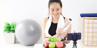 美丽的亚洲女人健康的博主展示了苹果水果和干净的减肥食品。在智能手机前录制视频直播在家。在线社交媒体上的健身影响者。