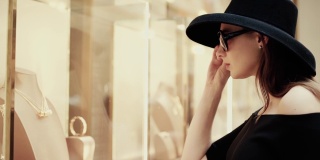 一个漂亮时髦的富家女孩戴着一顶帽子摘下眼镜，在一家奢侈品精品店的橱窗里看商品。慢动作