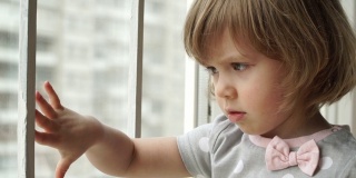 严肃的小女婴坐在窗台上，望着大城市的窗外，手梦幻般地滑在玻璃上。沉思的孩子注视着窗外的街道。