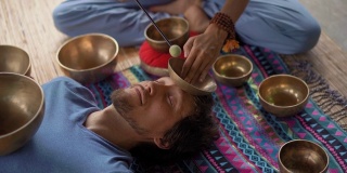 一名精通亚洲圣药的女子为一名年轻男子执行藏碗治疗仪式的超慢镜头。用西藏唱碗冥想。他们在一个凉亭里冥想，背景是美丽的瀑布