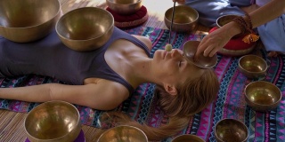 一名精通亚洲圣药的女子为一位年轻女性客户表演藏式碗治疗仪式的超慢镜头。用西藏唱碗冥想。他们在一个凉亭里冥想，背景是美丽的瀑布