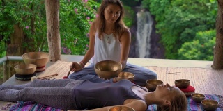 一名精通亚洲圣药的女子为一位年轻女性客户表演藏式碗治疗仪式的超慢镜头。用西藏唱碗冥想。他们在一个凉亭里冥想，背景是美丽的瀑布