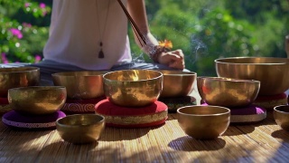 一名亚洲神药女大师表演藏式碗疗愈仪式的超慢镜头。用西藏唱碗冥想。她坐在凉亭里冥想，背景是美丽的瀑布视频素材模板下载