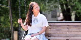 阳光明媚的日子里，可爱的中国年轻女子穿着日本学生风格的连衣裙在公园的绳梯上摇摆，戴着眼镜的漂亮女孩享受夏天的时光，超慢的动作，背光镜头。