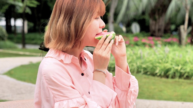 夏天女人吃西瓜片。微笑的中国女孩在公园吃西瓜。假期的生活方式。夏季的概念。深褐色的女人吃新鲜的西瓜，甜美的夏天。4k镜头，慢动作。