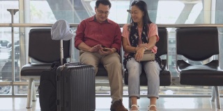 幸福的亚洲老年夫妇坐在候机楼的长椅上用智能手机等待起飞。老年人的旅程。老夫老妻。旅行。飞行。退休了。在一起。假期