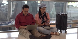 幸福的亚洲老年夫妇坐在地板上使用笔记本电脑和智能手机在候机楼机场等待离开。老年人的旅程。老夫老妻。旅行。飞行。退休