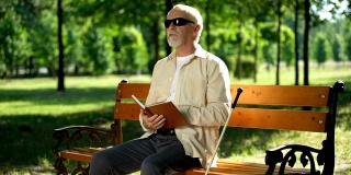 一位看盲文书的盲人，坐在夏日公园的长椅上休息