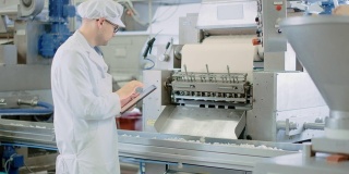 年轻男性质量主管或食品技术员正在检查饺子工厂的自动化生产。7 .员工使用平板电脑工作。他戴着白色卫生帽和工作袍。