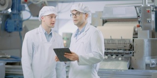 两个年轻的食品工厂员工讨论与工作有关的事情。男性技术员或质量经理使用平板电脑工作。他们戴着白色的卫生帽和工作袍。