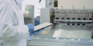 年轻的质量主管或食品技术员正在检查食品工厂的自动化生产。近距离拍摄的一个员工使用平板电脑工作。他戴着乳胶手套输入数据。