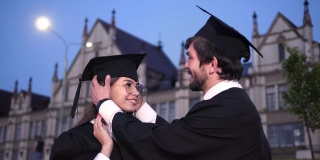 两个快乐的毕业生。一名男研究生帮女孩把帽子拉直，女孩朝他微笑