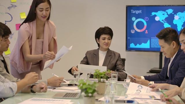 有魅力的亚洲女性在公司会议上展示工作计划、商业报告、给她的团队文件，不同的团队工作人员在办公室简报桌前做文书工作。