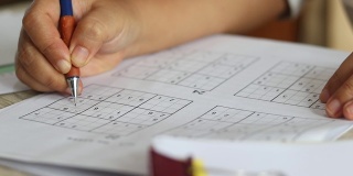 高级妇女的手试图解决数独谜与铅笔作为爱好在木制办公桌上。玩家将数字插入由9个方格组成的方格中，方格又细分为9个更小的方格。