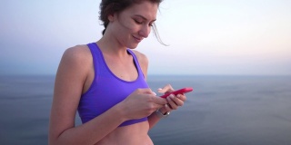 一个白人女运动员在社交网络上回复了很多信息，手里拿着手机。玩手机的时候玩运动，女人笑着看短信。日落在海边的山上