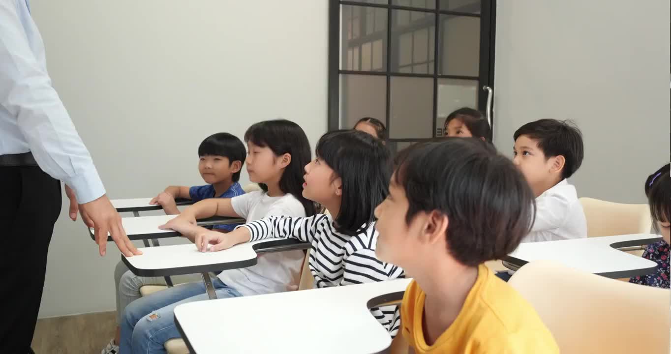 亚洲老师在学校的教室里教孩子们。小男孩和小女孩一起举手接受评论。教育理念、经验学习和技能发展。