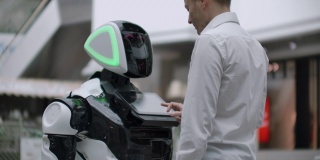 一名身穿衬衫的男子与一个白色机器人交流，机器人问问题，并用手指按屏幕。